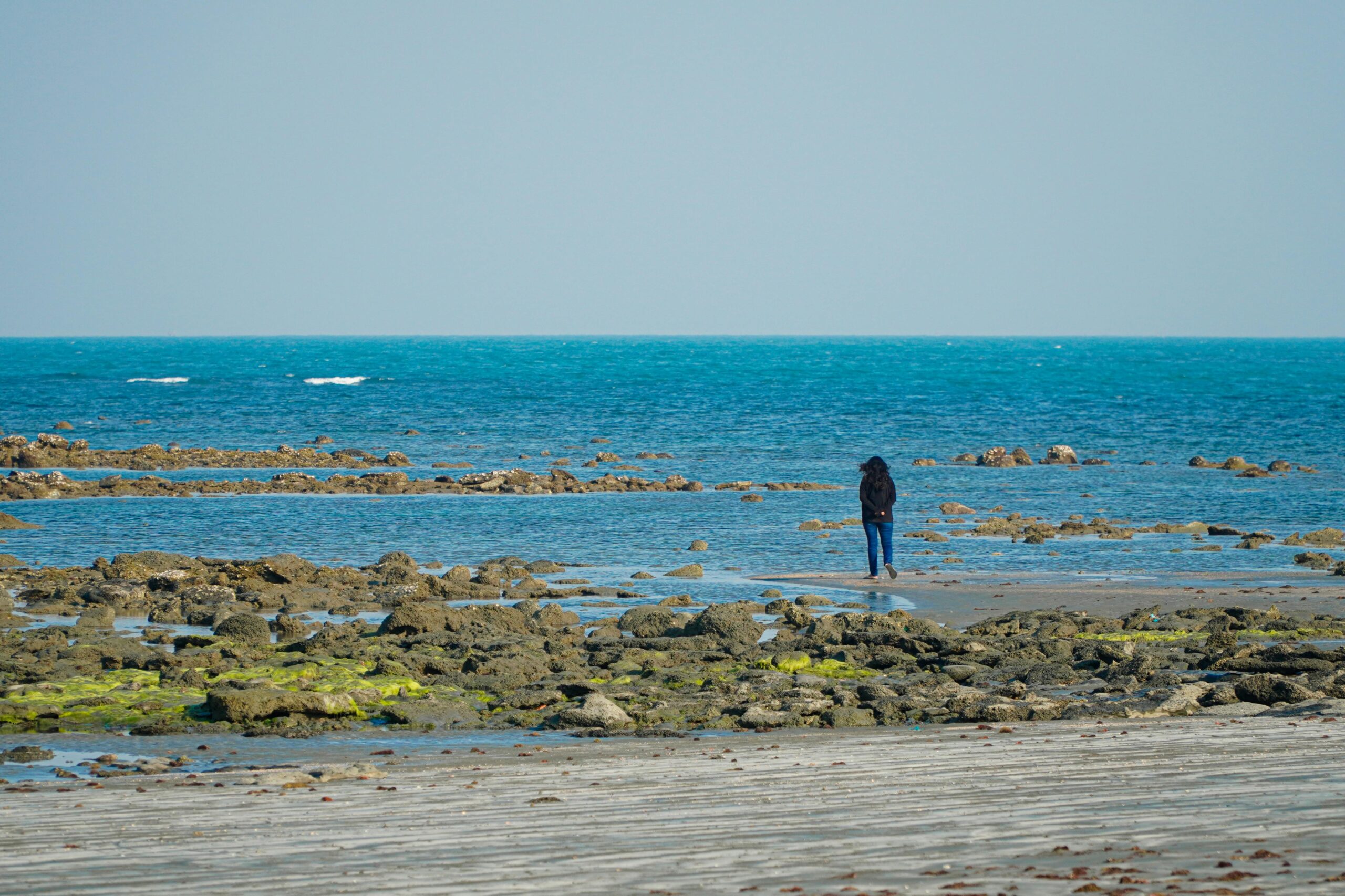 A solitary person walking along a tranquil, rocky beach with clear blue waters.